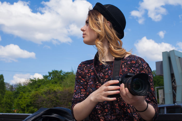 Young woman with digital camera looking away against sky