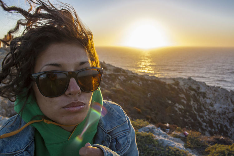 Young woman wearing sunglasses standing on cliff by sea during sunset