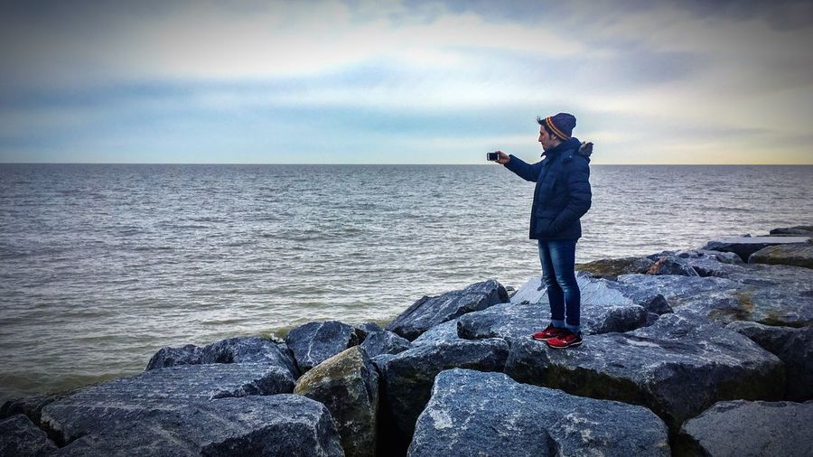 Full length of man photographing on rock by sea while using smart phone