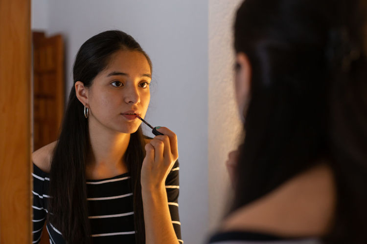 Young ethnic lady looking in mirror while applying lip gloss