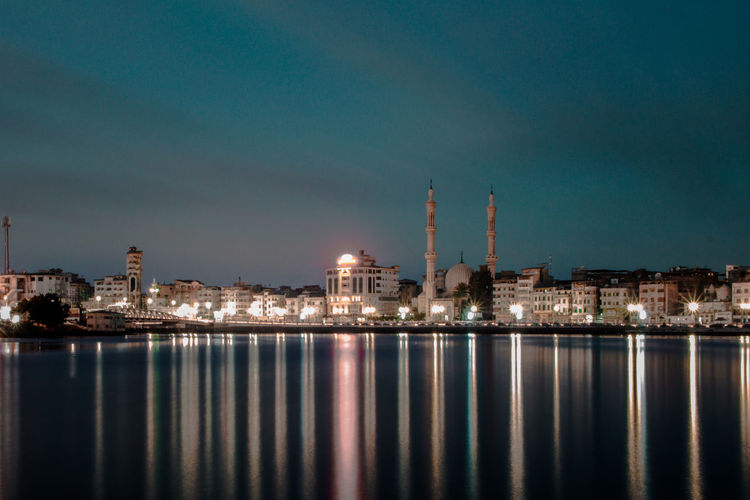 Illuminated city by river against sky at night