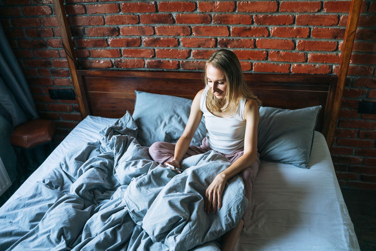 Adult woman with blonde long hair in pajamas using mobile phone sitting on bed in loft room at home