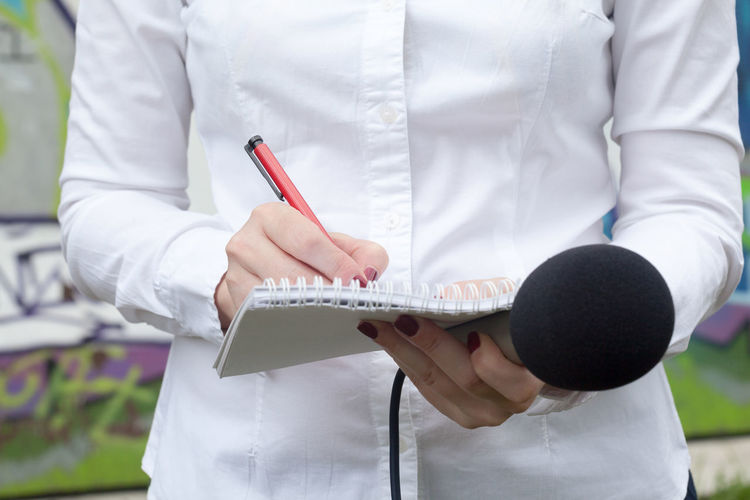 Midsection of female reporter holding microphone while writing on notepad