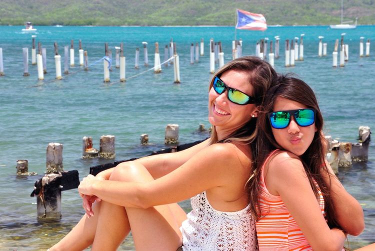 Portrait of female friends wearing sunglasses while sitting back to back at beach