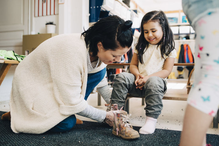 Side view of teacher helping happy girl wearing shoes in cloakroom at preschool