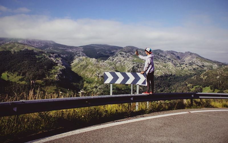 Man photographing mountains through mobile phone while standing on railing by road