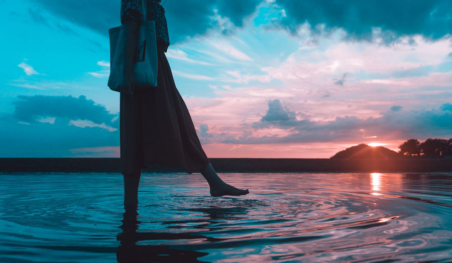 Low section of woman walking in sea against sky during sunset