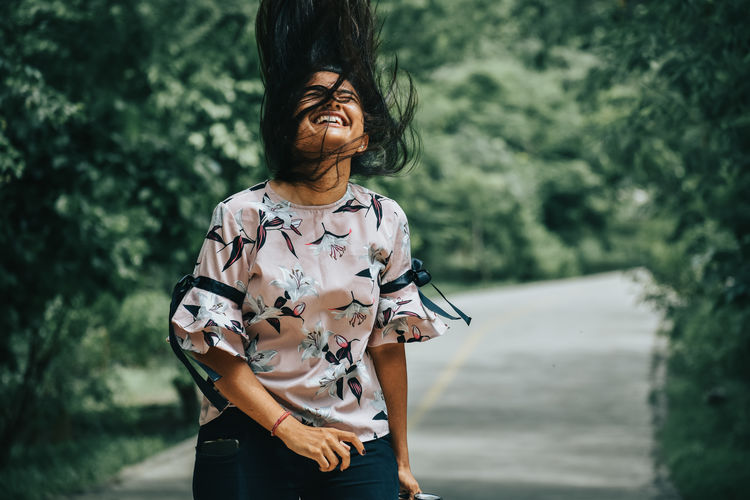 Smiling woman tossing hair while standing against trees