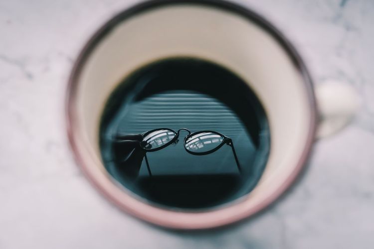 Cropped hand of person holding eyeglasses reflecting in coffee cup