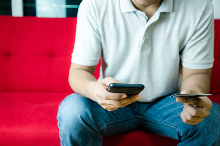 Midsection of man using mobile phone and holding credit card while sitting on sofa