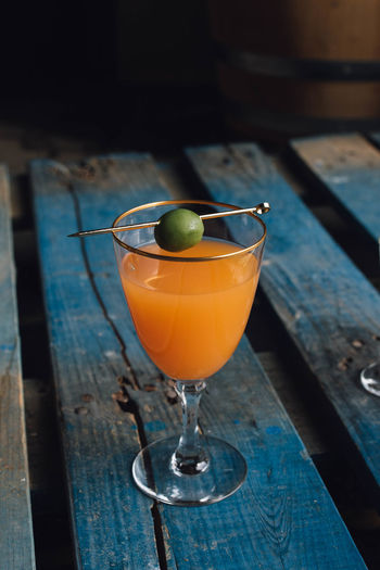 Carrot orange drink in wine glass with green olive on gold cocktail pick on blue wooden pallet 