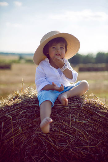Baby boy in straw hat and blue pants sitting on a haystack in a field in autumn