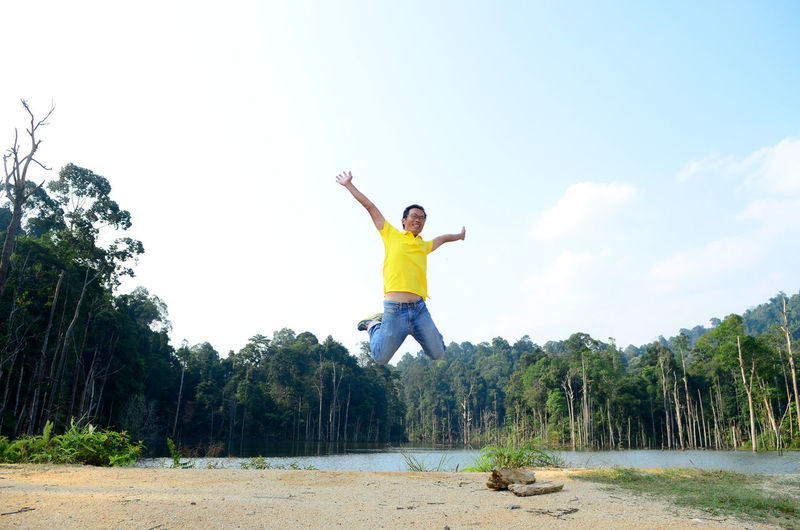 Cheerful man with arms outstretched jumping by lake against forest and sky