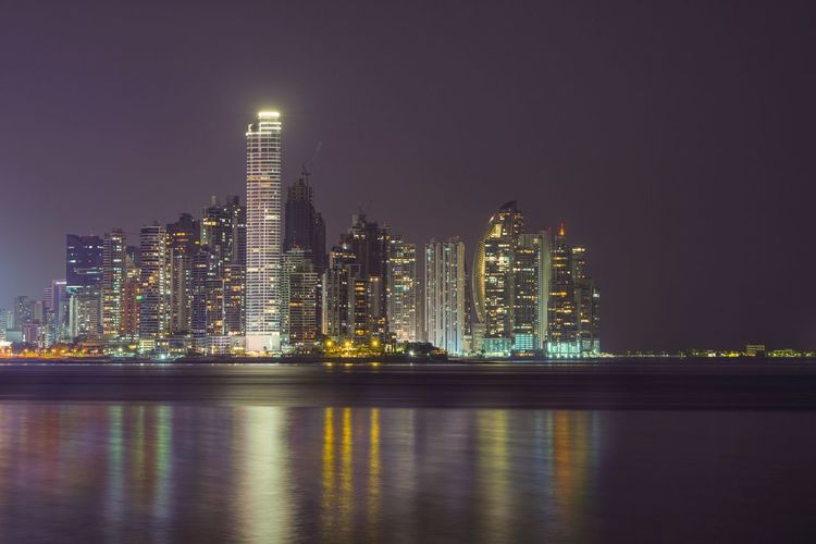 Illuminated modern buildings by river against sky at night