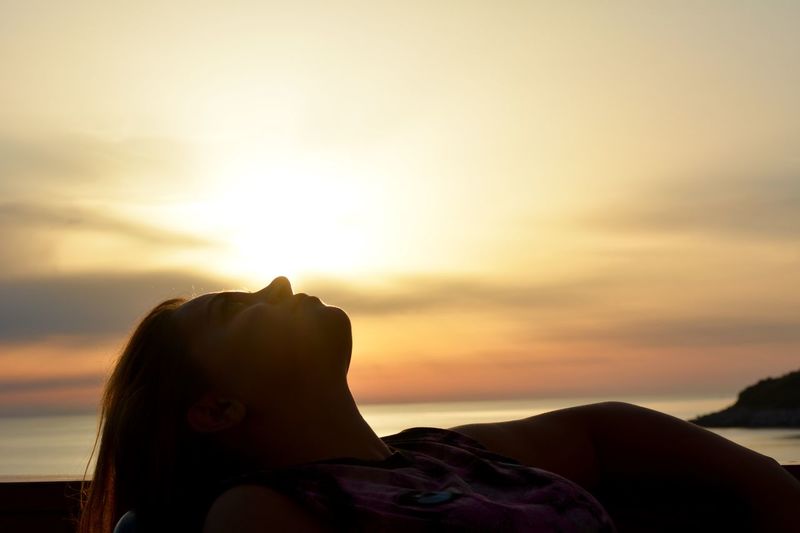 Side view of silhouette mid adult woman lying at beach against cloudy sky during sunset