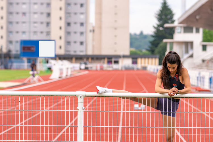 Young hispanic female athlete stretching legs near metal railing while warming up before running on stadium track