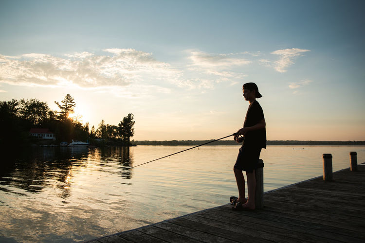 Adolescent boy fishing off dock on lake at sunset in ontario, canada.