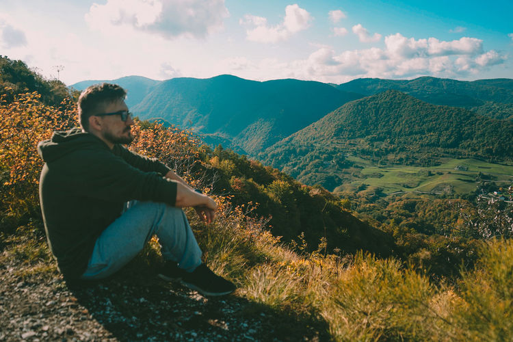 Young man sitting on mountain against sky