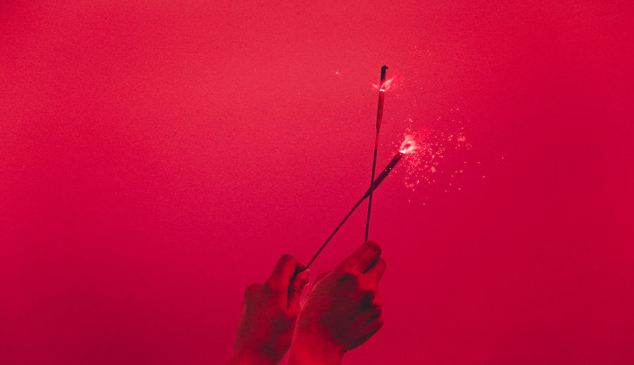 Cropped hand of person holding sparkler against red background