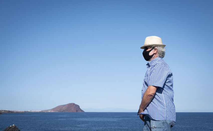 Senior man wearing mask standing by sea against clear sky