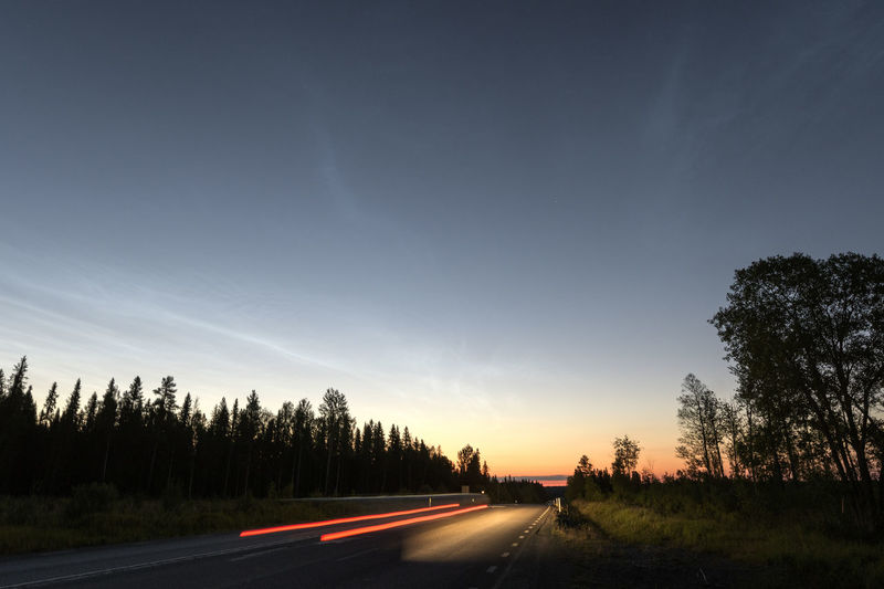 Road by trees against sky during sunset with noctilucent clouds 