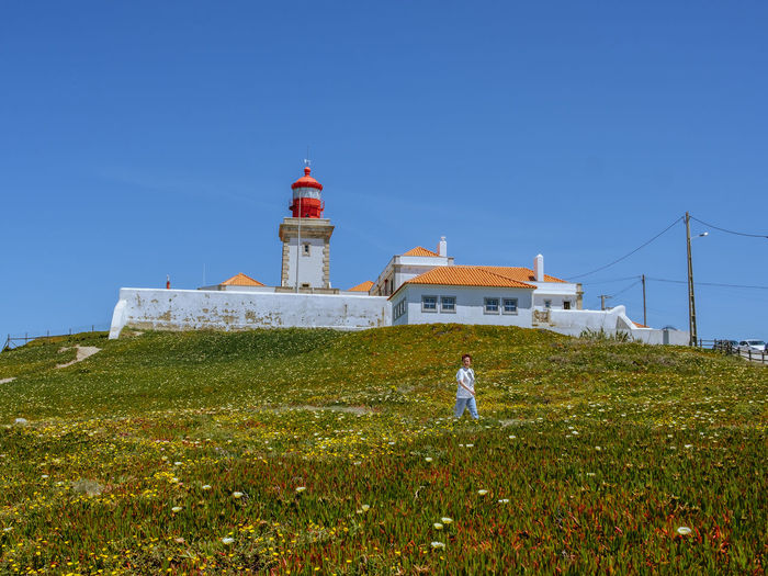 Lighthouse on field by building against clear blue sky