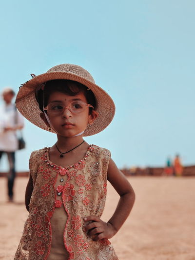 Cute girl wearing hat and eyeglasses looking away while standing at beach against sky