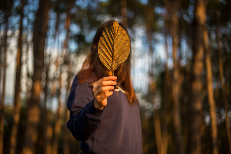 Woman covering face with leaf in forest during sunset