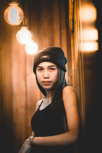 Portrait of young woman wearing hat standing against illuminated lamp