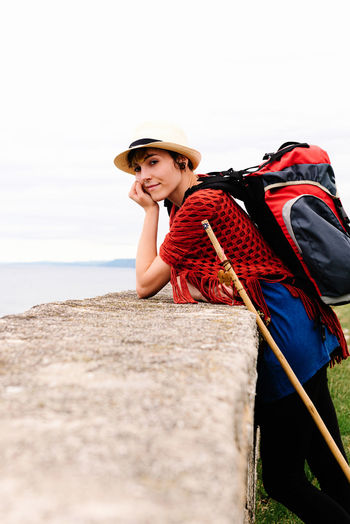 Side view of amazed young female pilgrim with backpack and trekking stick standing on sea coast looking at camera while enjoying trip along camino de santiago in spain