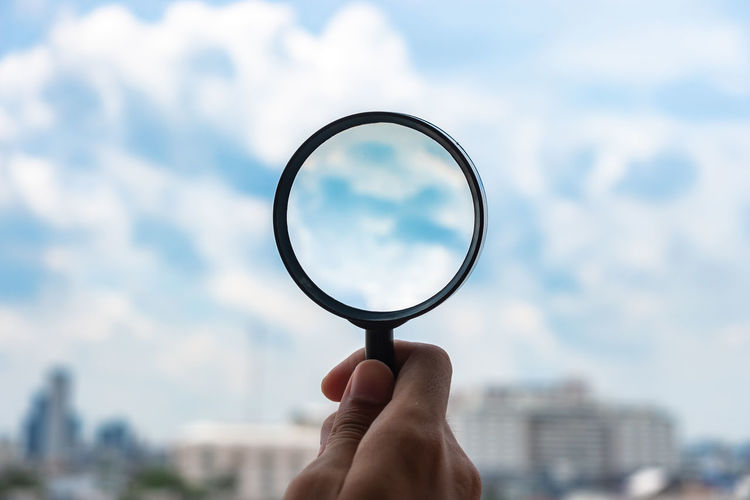 Cropped hand of person holding magnifying glass against sky