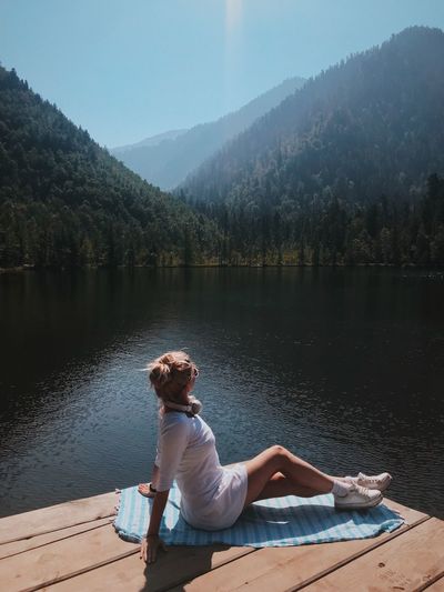 Full length of woman sitting on pier at lake against mountains