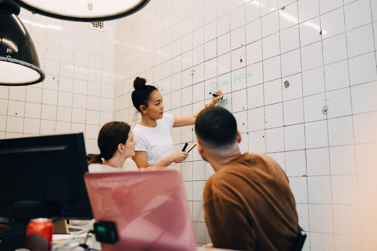 Young businesswoman brainstorming colleagues while writing strategy on tile wall at creative office