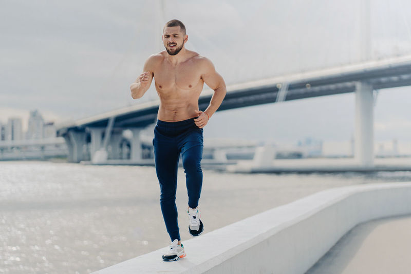 Full length of shirtless man exercising on retaining wall by river against bridge