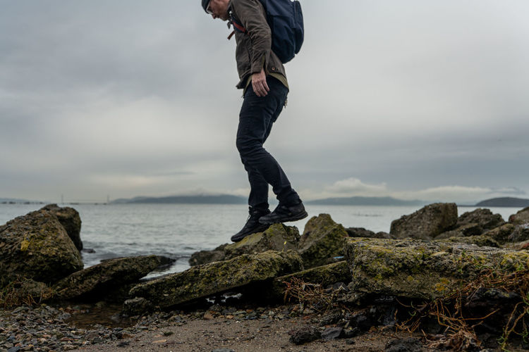Man with backpack climbs small path of rocks beside bay under gray sky