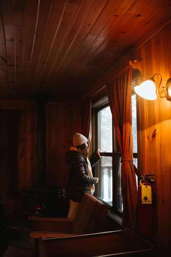 Side view of woman in outerwear and hat admiring snowy countryside while standing near window of dim wooden hut