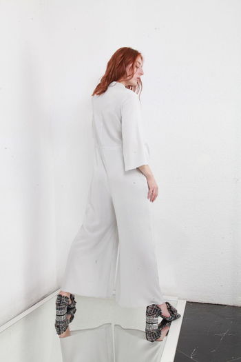 Back side view of fashion catalog style portrait of ginger woman in jumpsuit standing white wall