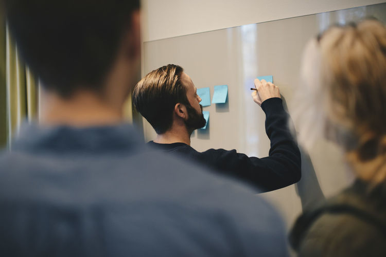 Businessman writing on adhesive note stuck to whiteboard with colleagues in foreground