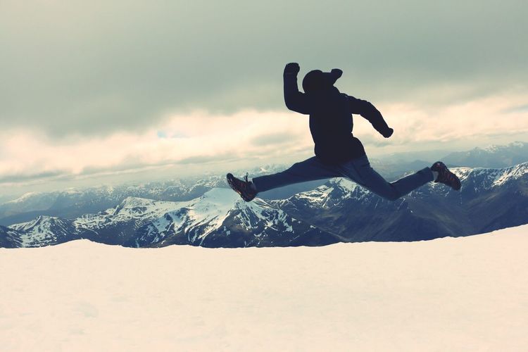 Full length of person jumping in mid-air on snowcapped mountain against cloudy sky