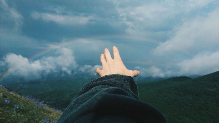 Close-up of hand pointing towards rainbow over countryside landscape