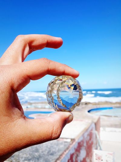 Cropped hand holding crystal ball against blue sky