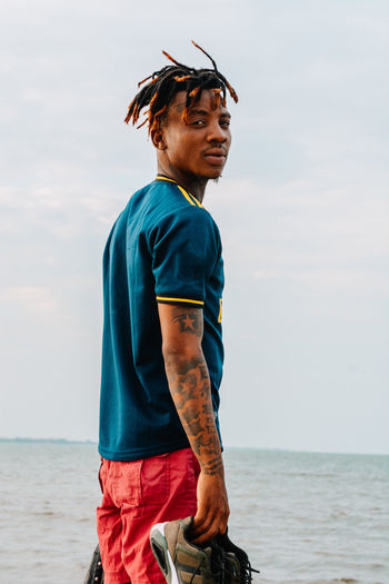 Young man looking at camera while standing in sea against sky