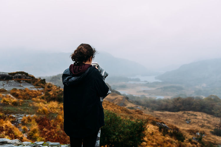 Rear view of teenage girl looking at mountains through telescope against sky