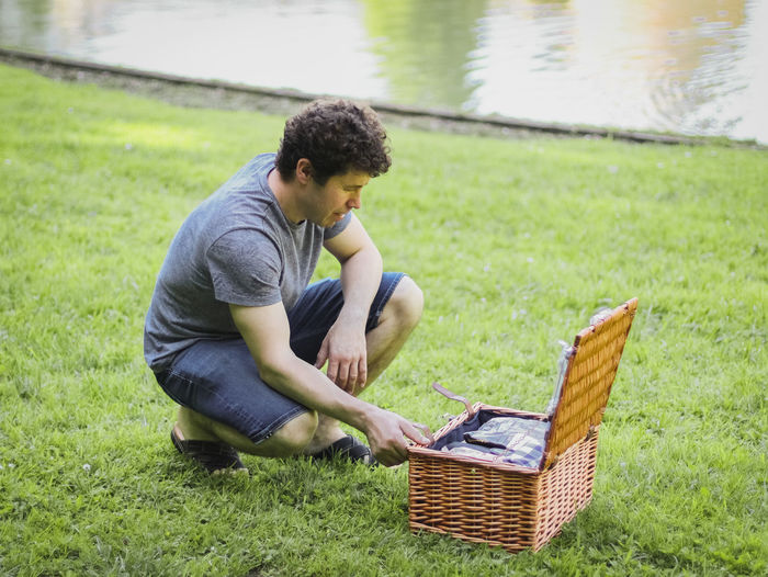 Handsome caucasian young male opens a wicker basket on a green meadow near a lake