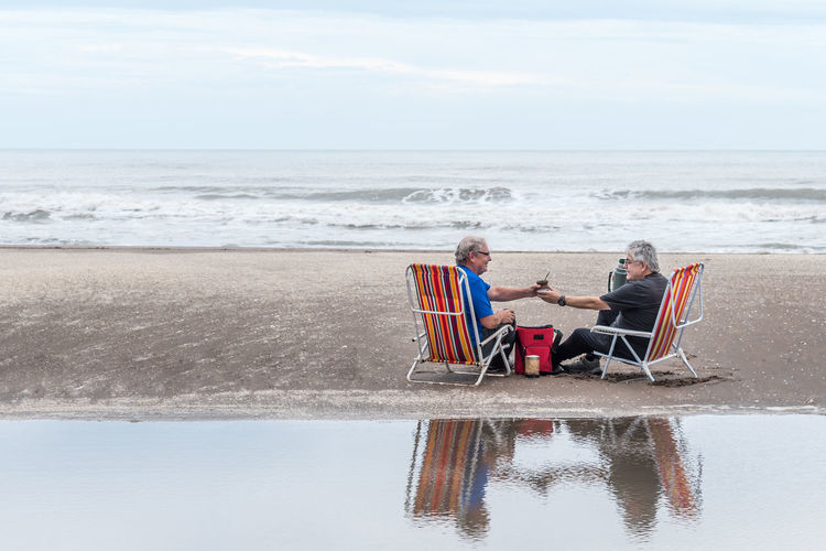 Two mature man with gray hair sitting on a beach chair drinking mate all reflected in the water. 