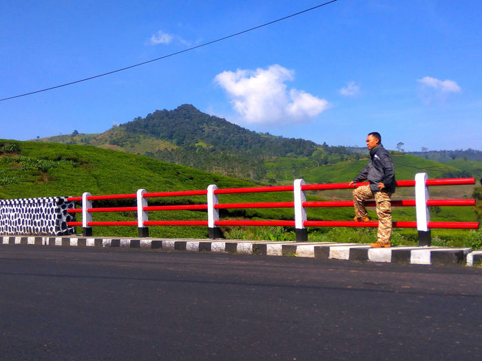 Man standing on railing by road against mountain and sky