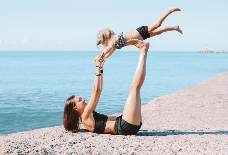 Mother with daughter exercising at sea shore against sky