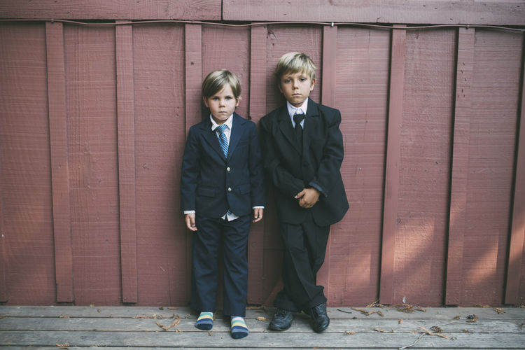 Portrait of confident brothers wearing suits while standing against wooden wall
