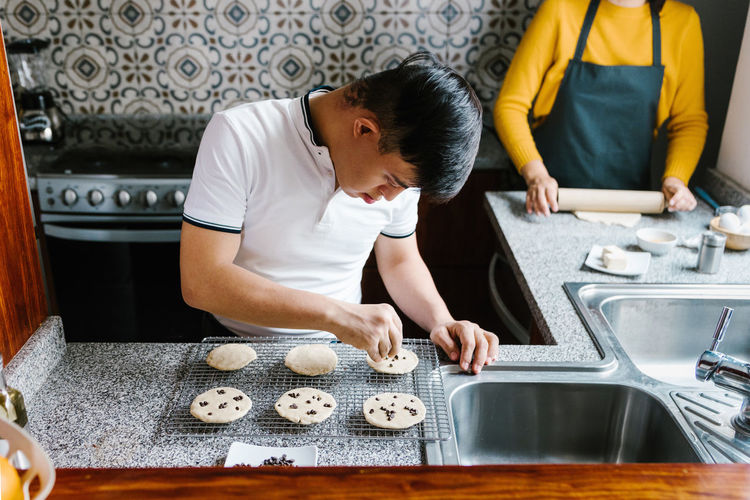 High angle of latin teenage boy with down syndrome decorating raw cookies with chocolate chips while cooking in kitchen at home