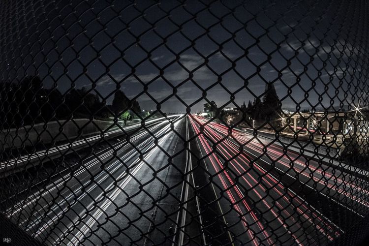 Light trails on road seen through chainlink fence at night